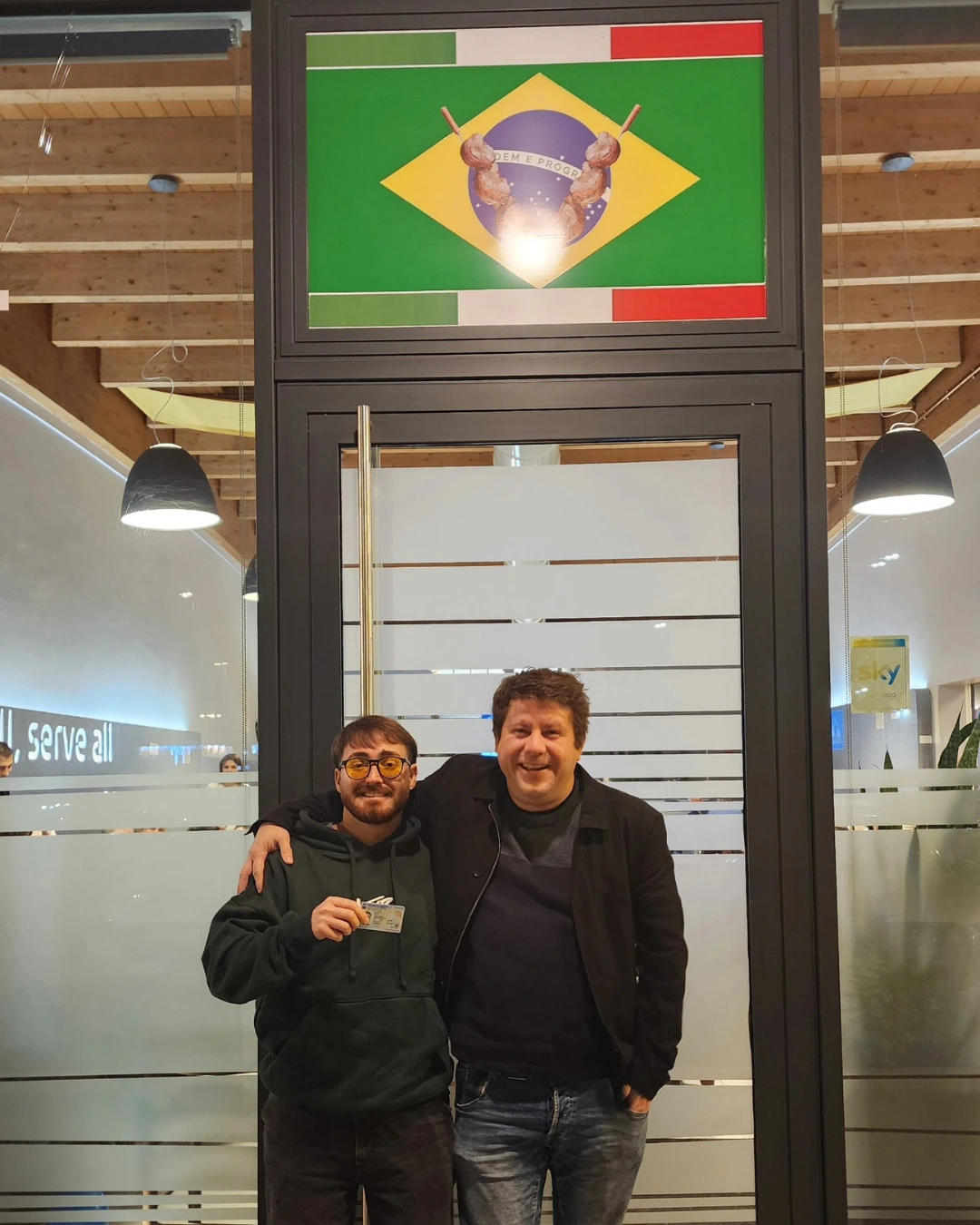 Dois clientes da Speranza posando em frente a um letreiro com as bandeiras do Brasil e da Itália